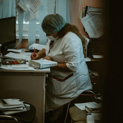 Healthcare worker wearing scrubs and face mask writing in an office setting, surrounded by documents.